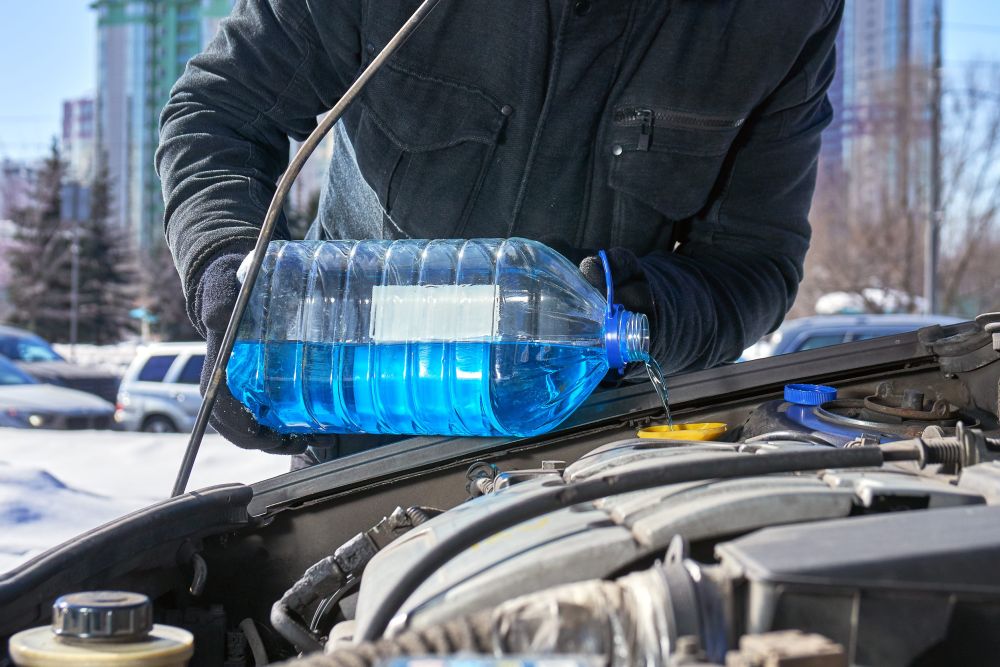 Vozač u tamnoj jakni sipa plavu tečnost iz velikog plastičnog kanistera u otvoreni motorni prostor automobila