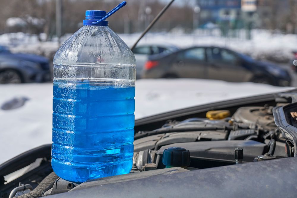 Velika plastična boca sa plavom rashladnom tečnošću postavljena pored otvorenog motornog prostora automobila tokom zime