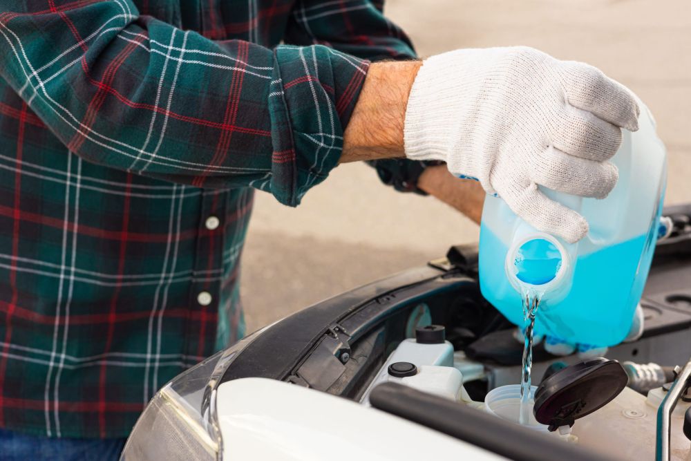 Osoba u belim zaštitnim rukavicama sipa plavu tečnost iz plastičnog kanistera u posudu ispod otvorene haube automobila